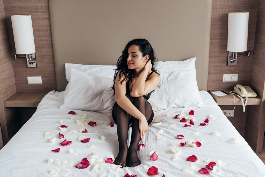 Sexy brunette girl in black stockings sitting on bed with rose petals.